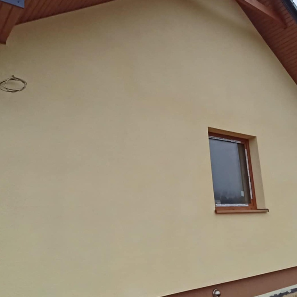 Fragment elewacji domu jednorodzinnego w kolorze kremowym, z brązową stolarką okienną i widocznym fragmentem dachu z drewnianą podbitką. Zewnętrzna lampa elewacyjna zamontowana pod oknem.