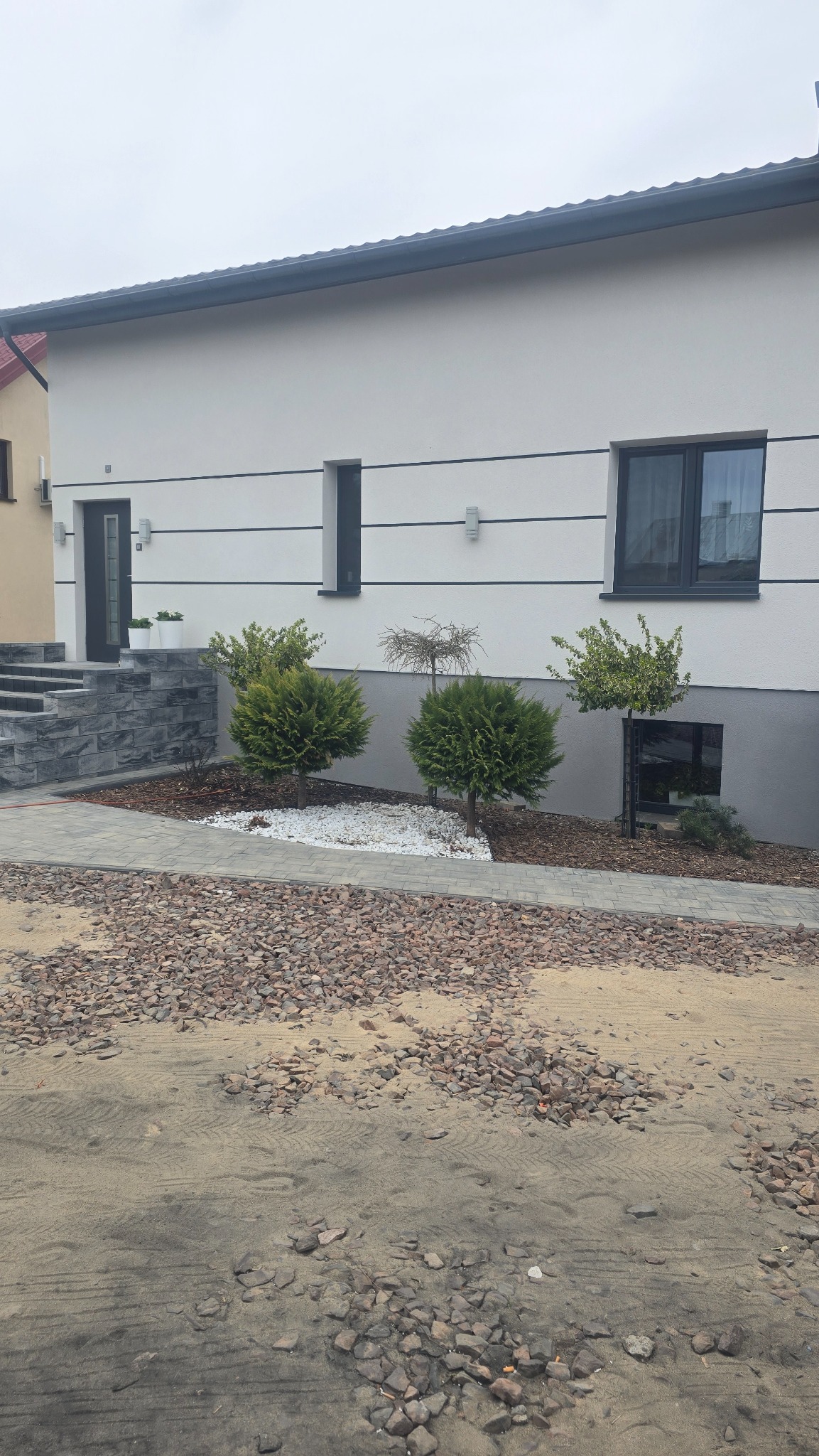 Wykończony dom z elewacją w odcieniach bieli i szarości, z elementami dekoracyjnymi w postaci pasów i oświetlenia. Uwagę zwraca zagospodarowanie terenu z roślinnością.