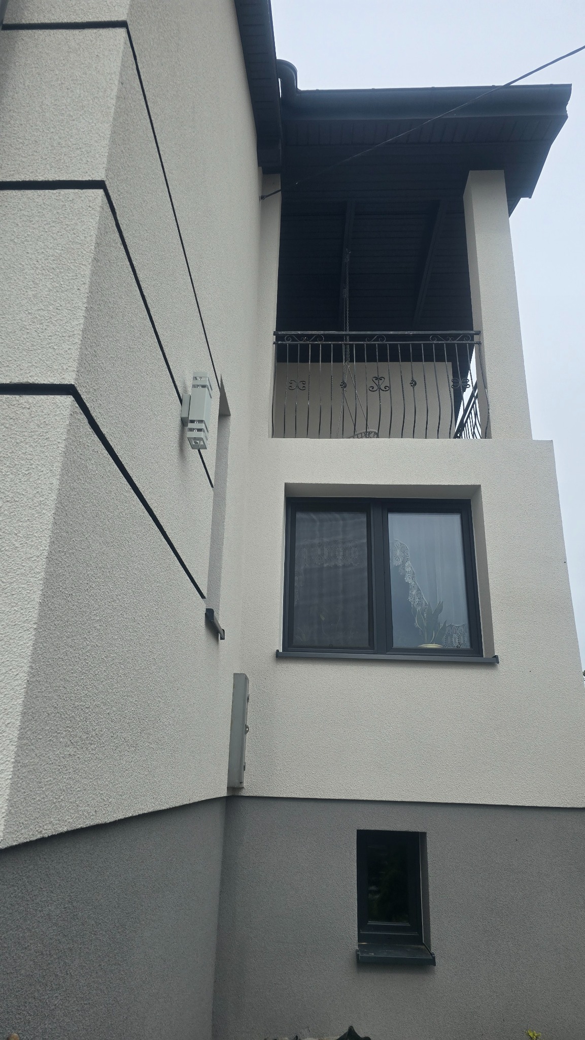 Elewacja dwukondygnacyjnego domu z balkonem i oknami. Jasny tynk z ciemnymi akcentami, nowoczesne oświetlenie zewnętrzne, ozdobna balustrada balkonu.