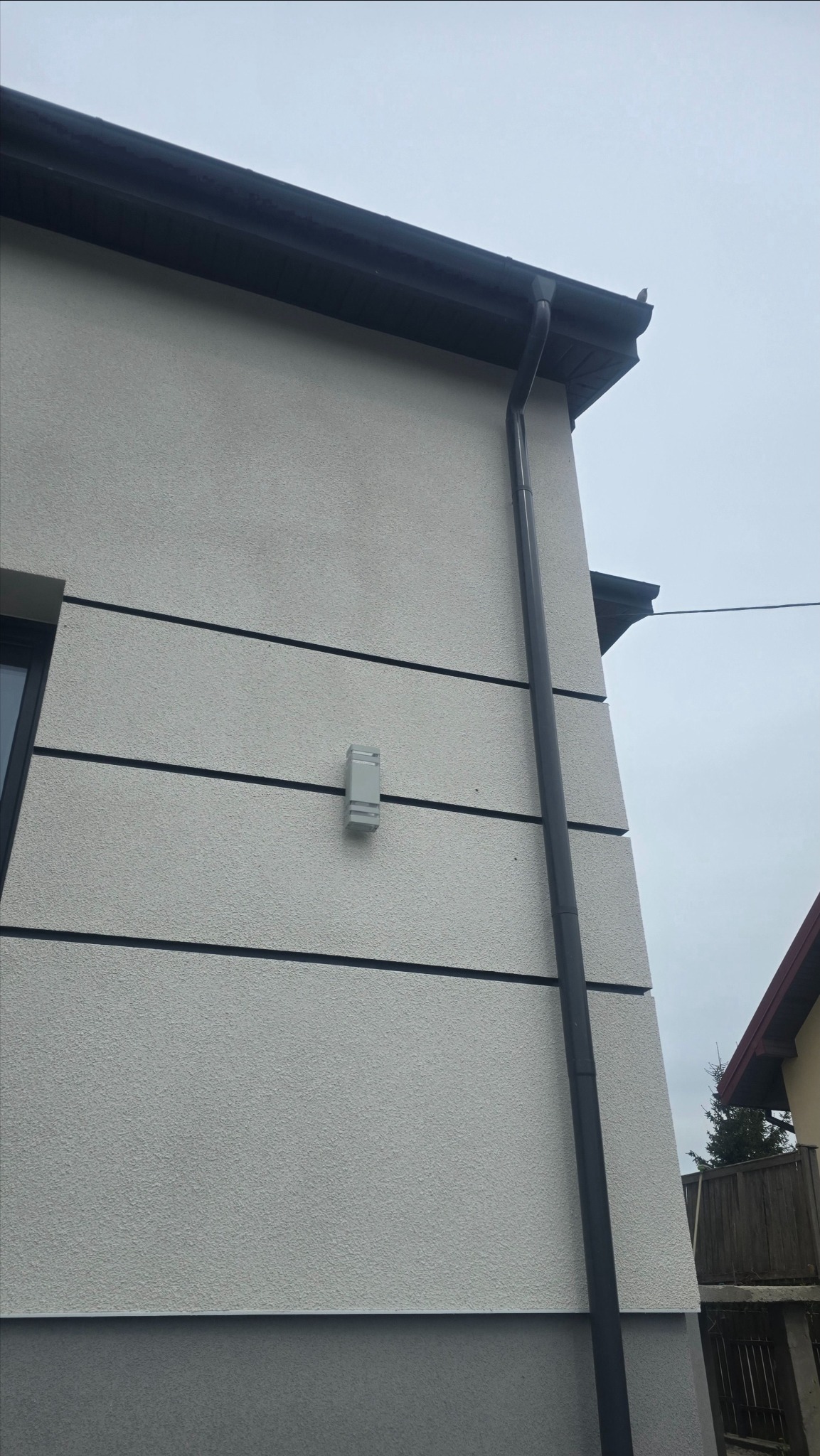 Fragment elewacji domu z szarym tynkiem strukturalnym, czarnymi akcentami i oświetleniem zewnętrznym. Widoczny róg budynku i rynna.