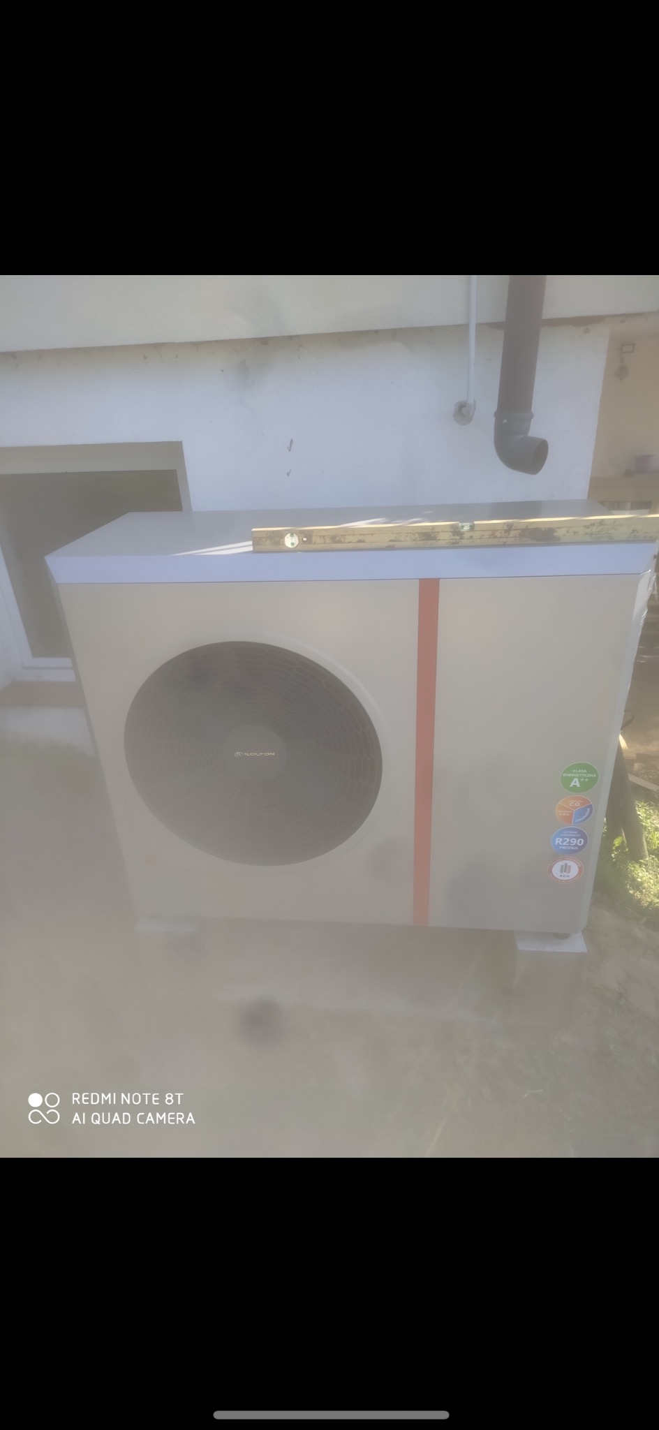 Zewnętrzna jednostka pompy ciepła z poziomnicą na górze, stojąca na betonowych bloczkach przy białej ścianie budynku z rurą spustową.