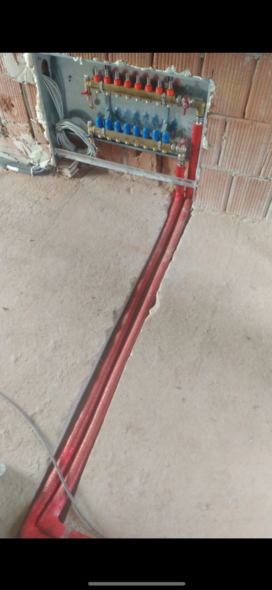 Rozdzielacz instalacji grzewczej z czerwonymi rurami w bruździe w podłodze, widok na ścianę z cegły i kable w puszce.
