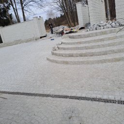 MET-BRUK Marcin Sałacki - Granitowa nawierzchnia z subtelnymi łukami i schodami, w tle pracownik z taczką. Widoczny drenaż liniowy i elementy muru z szarej kostki.