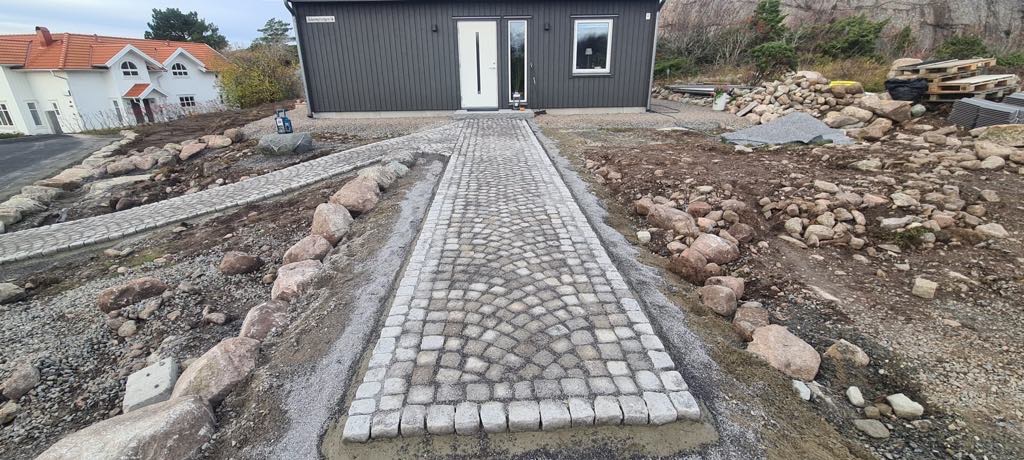 Granitowa kostka ułożona w łukowy wzór tworząca ścieżkę do ciemnoszarego domu z białymi drzwiami i oknem, otoczona dużymi kamieniami i przygotowanym podłożem.