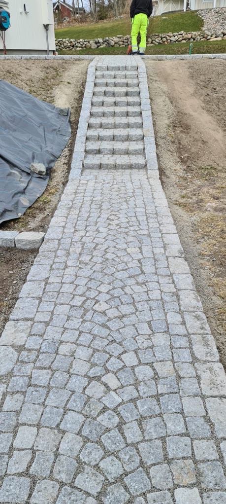Granitowa ścieżka z kostki prowadząca do schodów, ułożona w delikatny łuk, z pracownikiem w odblaskowej kurtce w tle.