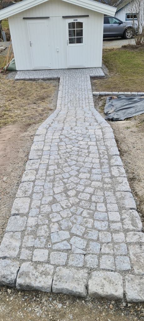 Ścieżka z jasnoszarej kostki granitowej prowadząca do białego domku narzędziowego, ułożona w łukowaty wzór, z widocznym samochodem w tle.