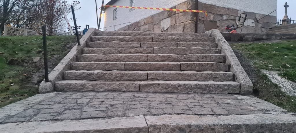Granitowe schody prowadzące do budynku z elementami zabezpieczeń budowlanych w tle, wykonane z nieregularnych, szarych bloków.