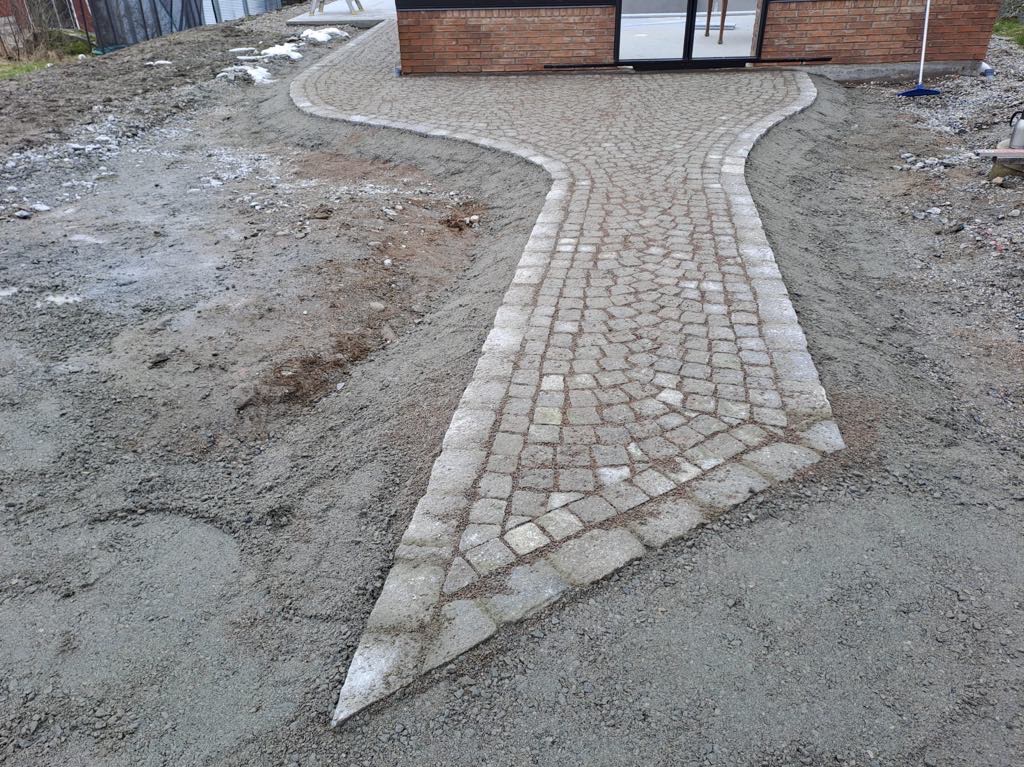 Ścieżka z kostki granitowej w Dzierżoniowie, prowadząca do budynku z cegły, otoczona świeżo przygotowanym podłożem.