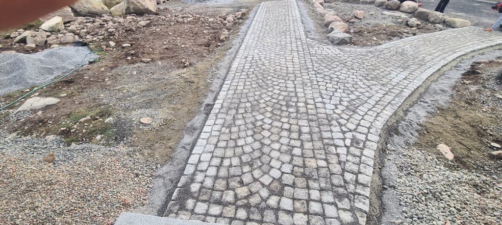 Granitowa ścieżka rozwidlająca się na dwie drogi, w trakcie budowy, z widocznym podsypem i kamieniami dekoracyjnymi po bokach.