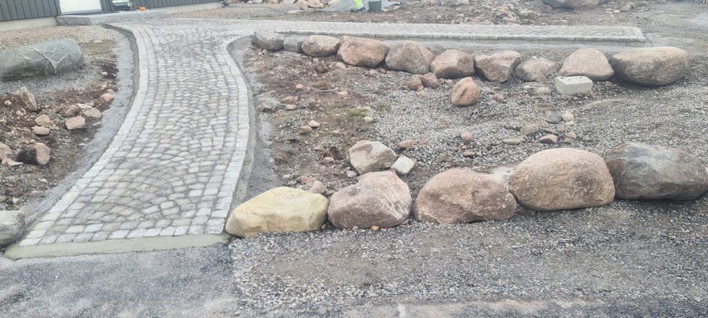 Ułożona ścieżka z kostki granitowej w kształcie wachlarza, obramowana betonem, z dużymi kamieniami po bokach, na żwirowym podłożu.