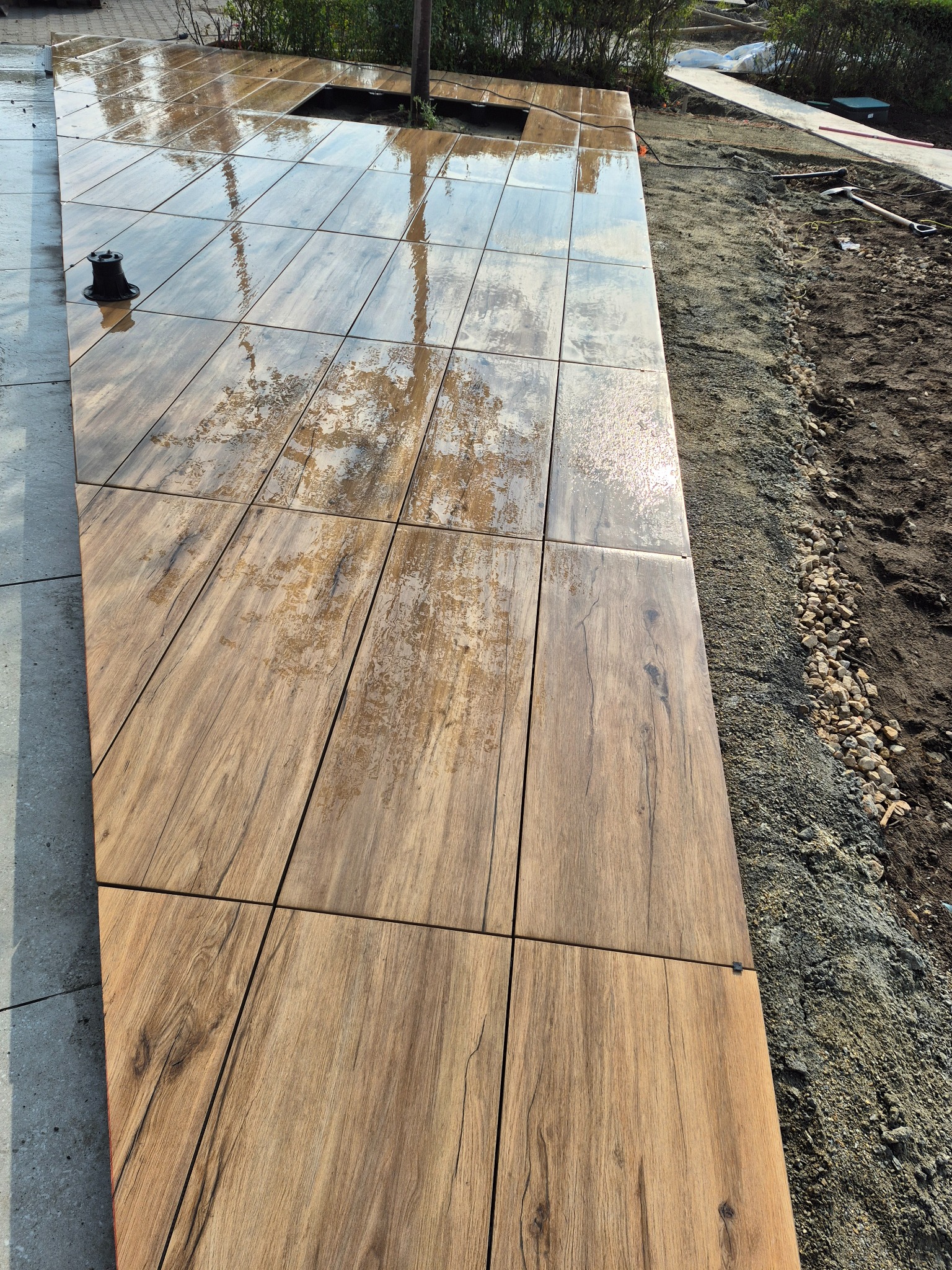 Ułożona, mokra, drewnopodobna kostka brukowa w kwadraty, z widoczną przestrzenią na roślinę. Obok ziemia i narzędzia. Perspektywa z góry.