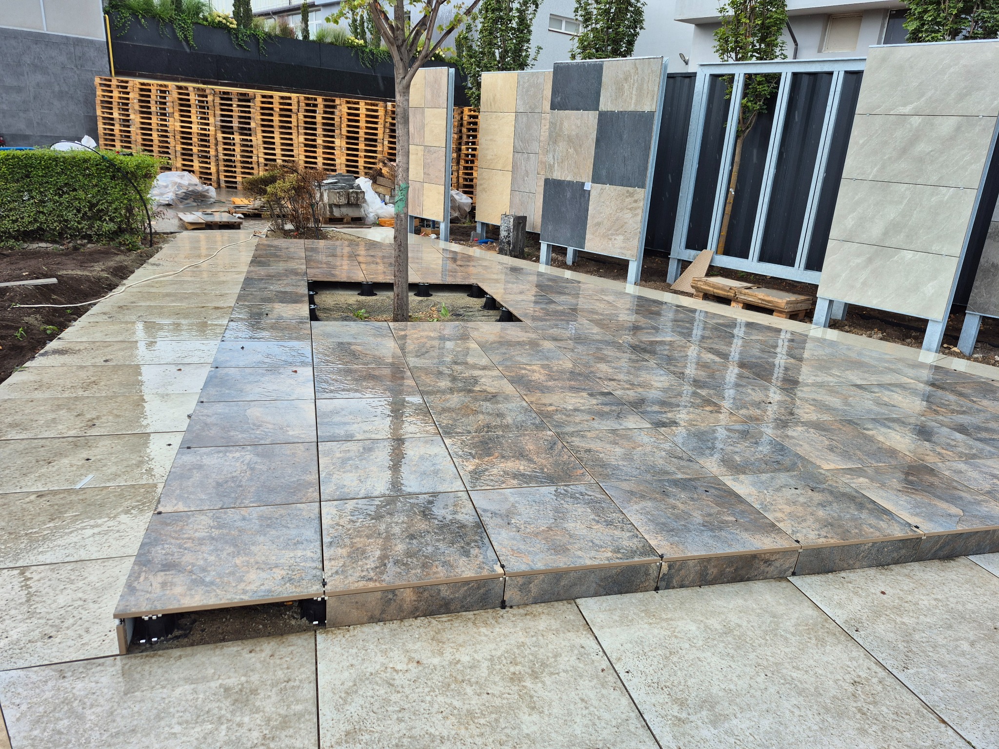 Ułożona, częściowo mokra, tarasowa nawierzchnia z płyt, z widocznymi elementami montażowymi i przygotowanymi otworami na rośliny. W tle ekspozycja wzorów.