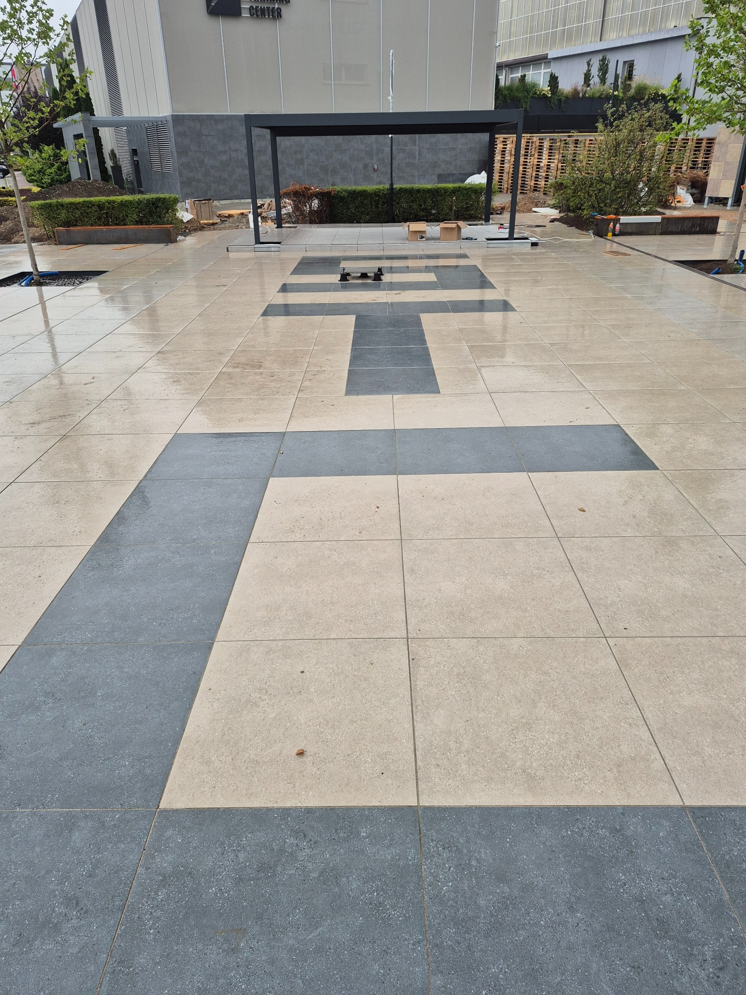 Nowa nawierzchnia z jasnych i ciemnych płyt w geometryczny wzór krzyża, na zewnątrz budynku z elementami zieleni i czarną pergolą w tle. Powierzchnia lekko mokra.