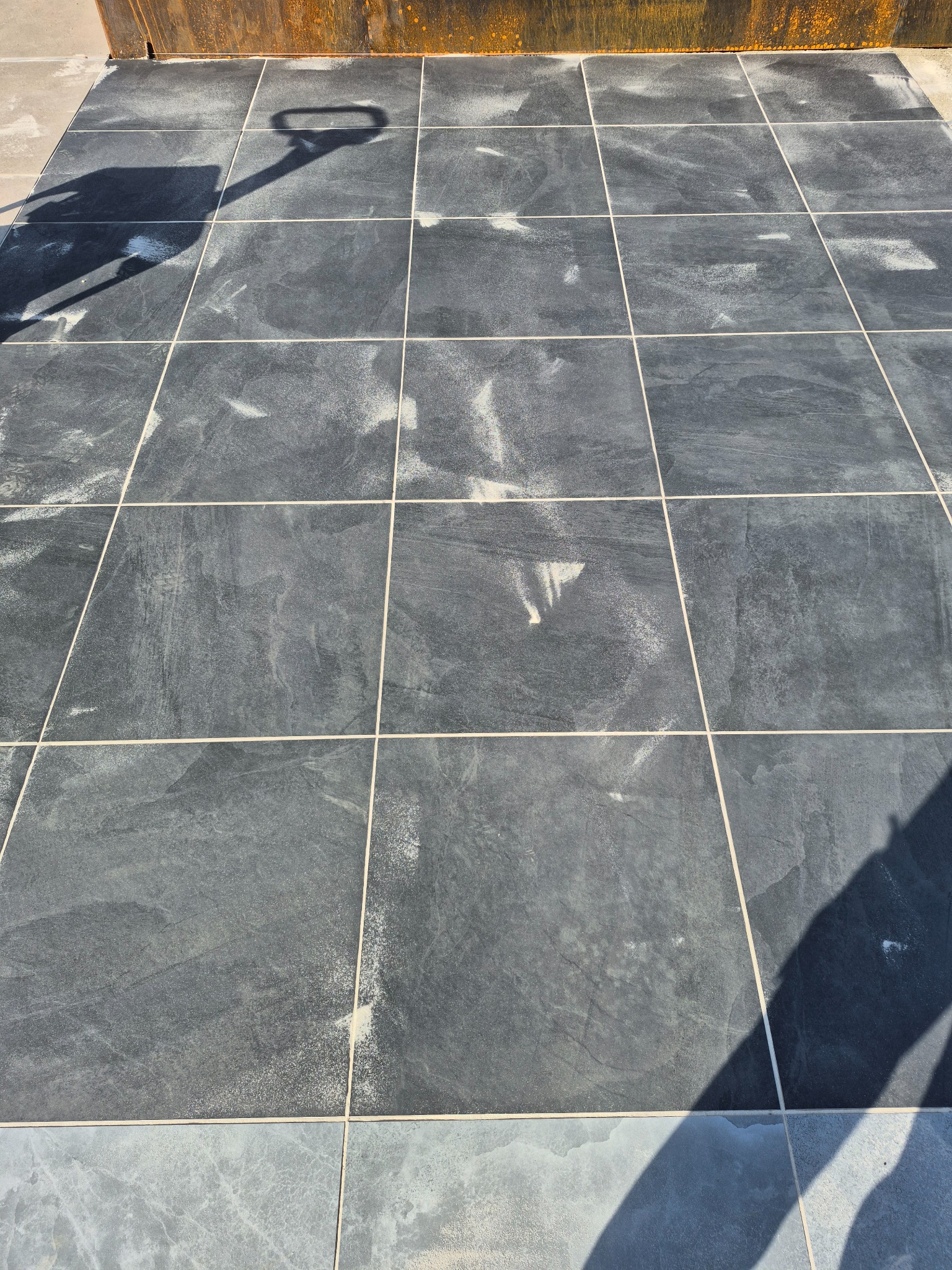 Szare, kwadratowe płyty ułożone na zewnątrz z jasnymi fugami. Na płytach widoczne ślady białego pyłu i cień osoby z łopatą.