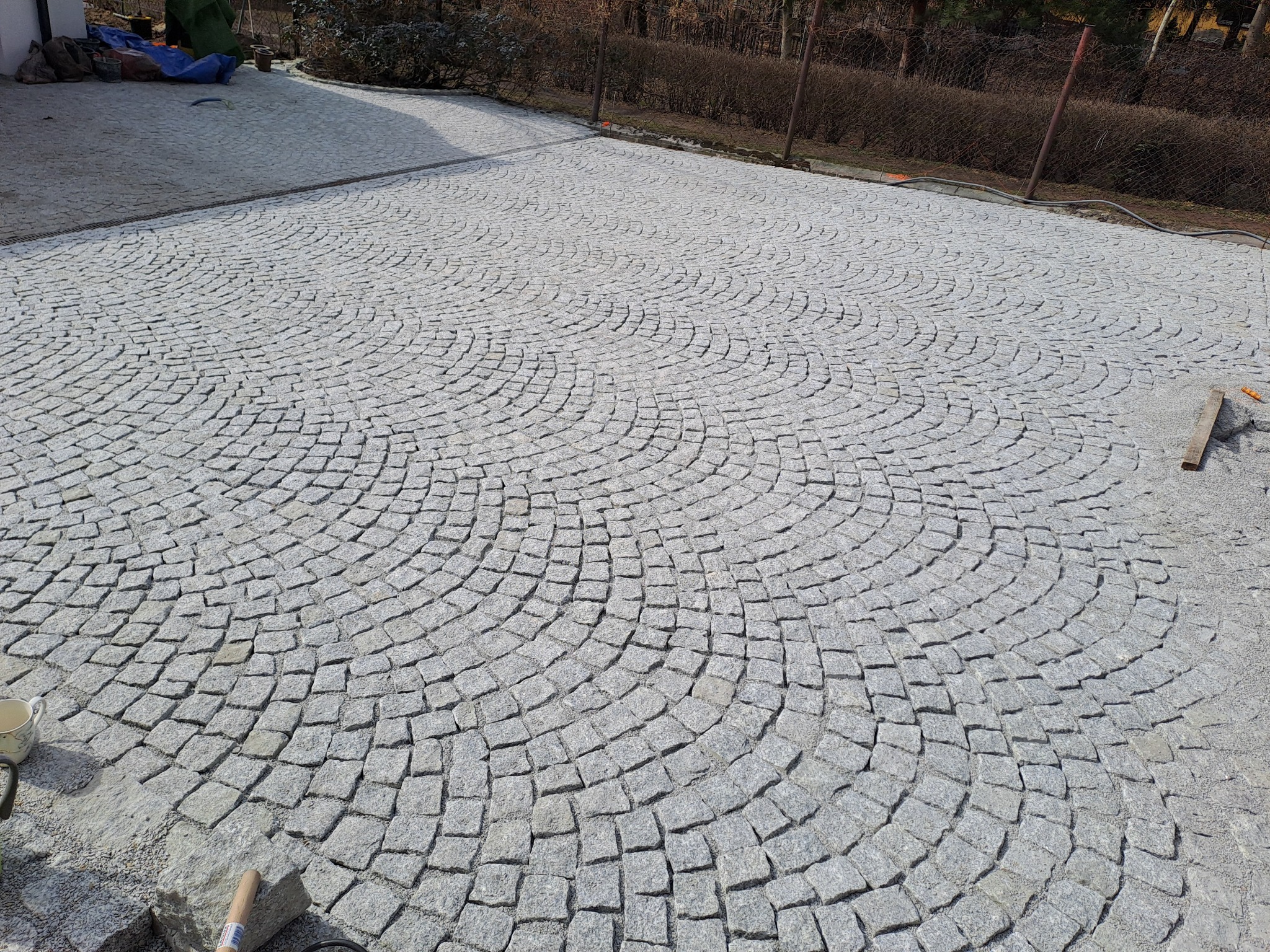 Ułożona granitowa kostka w kształcie wachlarza tworzy wzorzystą nawierzchnię podjazdu. Widoczne narzędzia brukarskie i surowce. Jasne, naturalne światło.