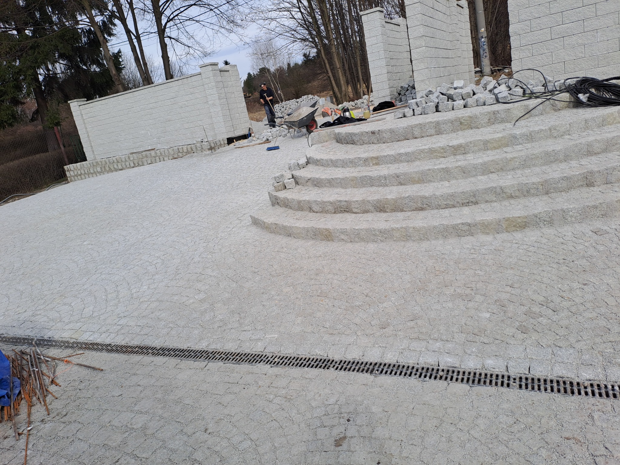 Granitowa nawierzchnia z subtelnymi łukami i schodami, w tle pracownik z taczką. Widoczny drenaż liniowy i elementy muru z szarej kostki.