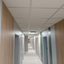 Długi korytarz biurowy w trakcie prac wykończeniowych, widoczne panele ścienne imitujące drewno, sufit podwieszany kasetonowy oraz wnęki drzwiowe w szarym kolorze.