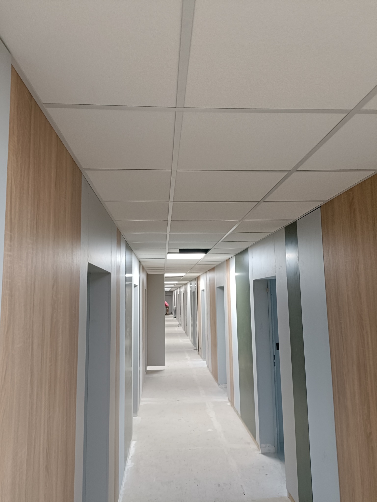 Długi korytarz biurowy w trakcie prac wykończeniowych, widoczne panele ścienne imitujące drewno, sufit podwieszany kasetonowy oraz wnęki drzwiowe w szarym kolorze.