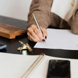 Osoba pisząca na kartce na biurku w Łodzi, widoczny drewniany keyboard, notatnik i telefon.