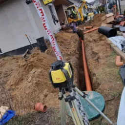 Wykonanie wykopu pod instalacje, widoczny laser niwelacyjny Leica na statywie, rury kanalizacyjne w wykopie, w tle koparka i budynek w stanie surowym.