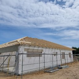 Furious-Bud - Budowa domu w trakcie realizacji: ściany z białych bloczków, drewniana konstrukcja dachu w trakcie montażu, rusztowania wokół budynku, błękitne niebo z chmurami w tle.