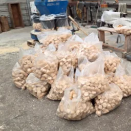 Stos przezroczystych worków wypełnionych drewnianymi pelletami, ustawionych na betonowej posadzce w hali produkcyjnej, z widocznym sprzętem do produkcji pelletu w tle.