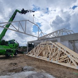 Furious-Bud - Montaż drewnianej więźby dachowej z użyciem dźwigu teleskopowego na placu budowy domu z białych bloczków, w tle widoczne drzewa i pochmurne niebo.