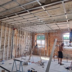 Furious-Bud - Montaż metalowej konstrukcji pod płyty gipsowo-kartonowe w pomieszczeniu z odsłoniętą cegłą, widoczne profile sufitowe i ścienne, dwaj pracownicy na drabinach, jeden bez koszulki.