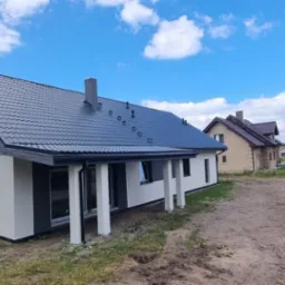 Nowoczesny dom z dwuspadowym dachem z blachy w kolorze grafitowym, biało-szara elewacja, widok z zewnątrz w słoneczny dzień, w tle inny dom i drzewa.