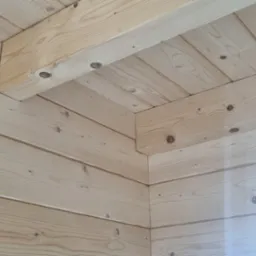 Wnętrze domu w trakcie budowy, widoczny fragment ściany i sufitu wykonane z jasnych, drewnianych desek łączonych na pióro i wpust, z widoczną izolacją z wełny mineralnej w narożniku.