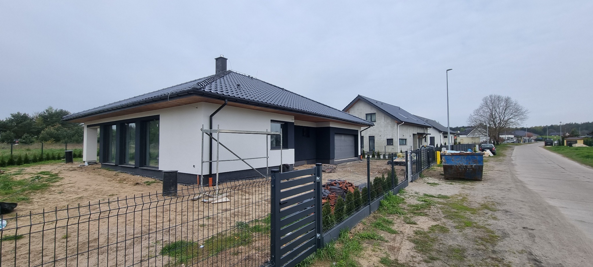 Nowoczesny dom jednorodzinny z czarnym dachem i elewacją w trakcie wykończenia, widoczne rusztowania, ogrodzenie panelowe i sąsiednie budynki w tle.