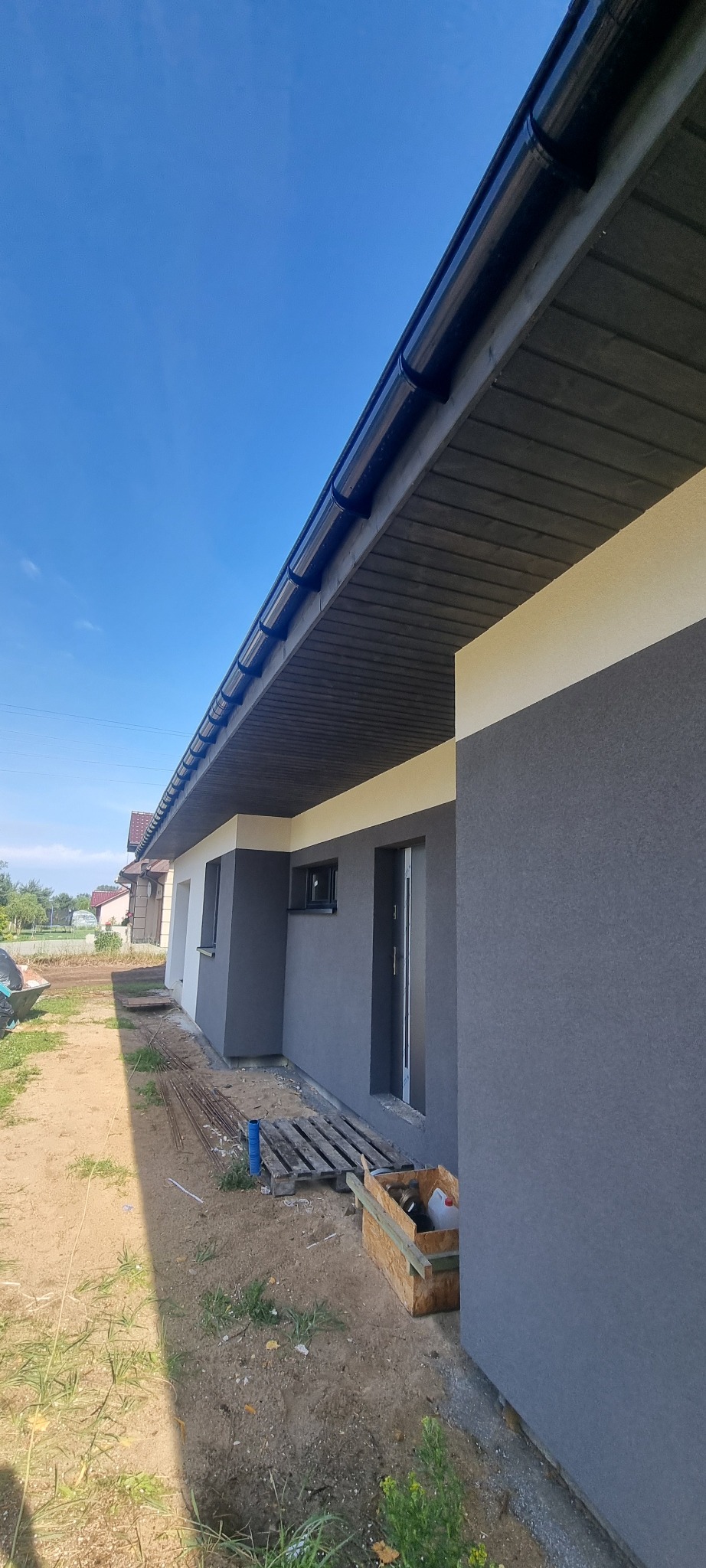 Fragment elewacji domu jednorodzinnego z widocznymi rynnami, oknami i drzwiami w odcieniach szarości, z paletą i narzędziami w tle, sugerującymi prace wykończeniowe.