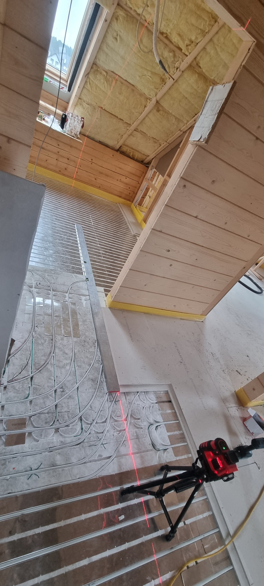 Wnętrze w trakcie budowy, widoczne ogrzewanie podłogowe, izolacja na poddaszu i laser krzyżowy na statywie, wyznaczający poziom na ścianie z boazerii.