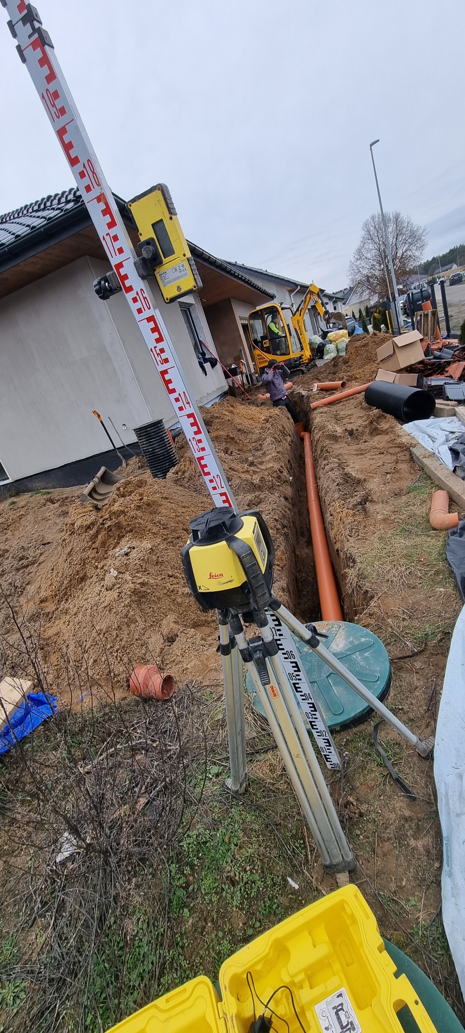 Wykonanie wykopu pod instalacje, widoczny laser niwelacyjny Leica na statywie, rury kanalizacyjne w wykopie, w tle koparka i budynek w stanie surowym.