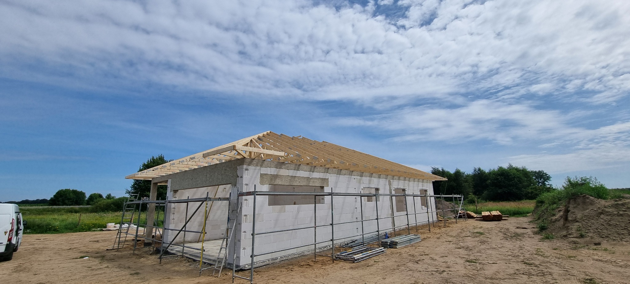 Budowa domu w trakcie realizacji: ściany z białych bloczków, drewniana konstrukcja dachu w trakcie montażu, rusztowania wokół budynku, błękitne niebo z chmurami w tle.