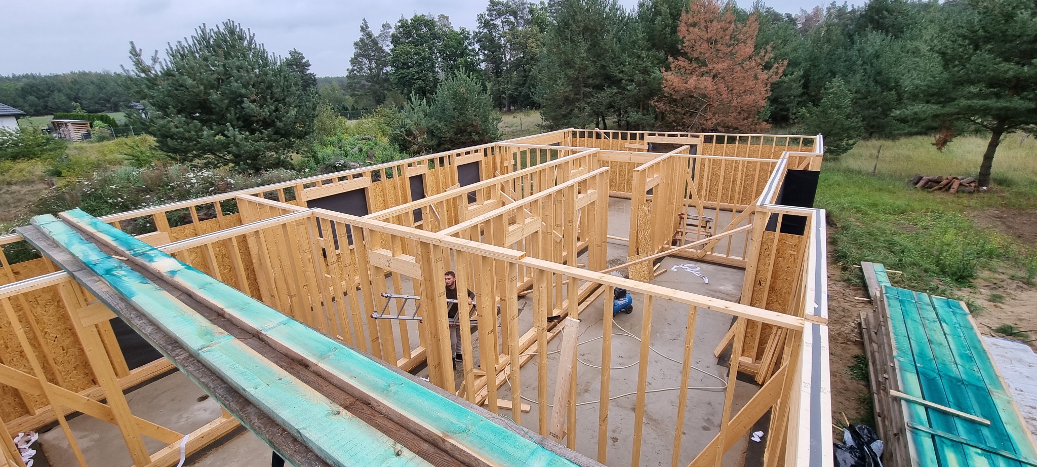 Budowa domu szkieletowego w Węgorzynie: widok z góry na drewniany szkielet konstrukcji z widocznym pracownikiem na drabinie wewnątrz, przygotowane deski w kolorze turkusowym leżące na zewnątrz...