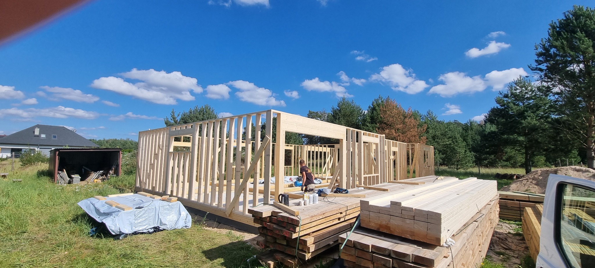 Wznoszenie drewnianej konstrukcji domu w technologii szkieletowej na fundamencie, widoczne deski i pracownik w trakcie montażu, błękitne niebo w tle.