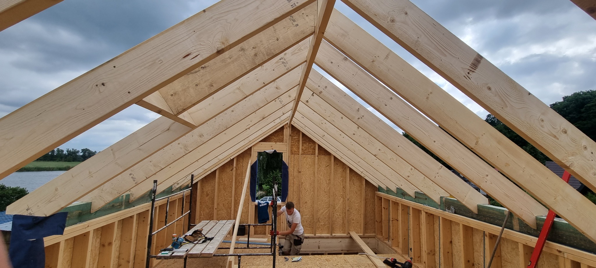 Wnętrze domu w trakcie budowy, widoczne drewniane elementy konstrukcyjne dachu i ścian, pracownik na rusztowaniu przy oknie, w tle krajobraz z jeziorem i drzewami.