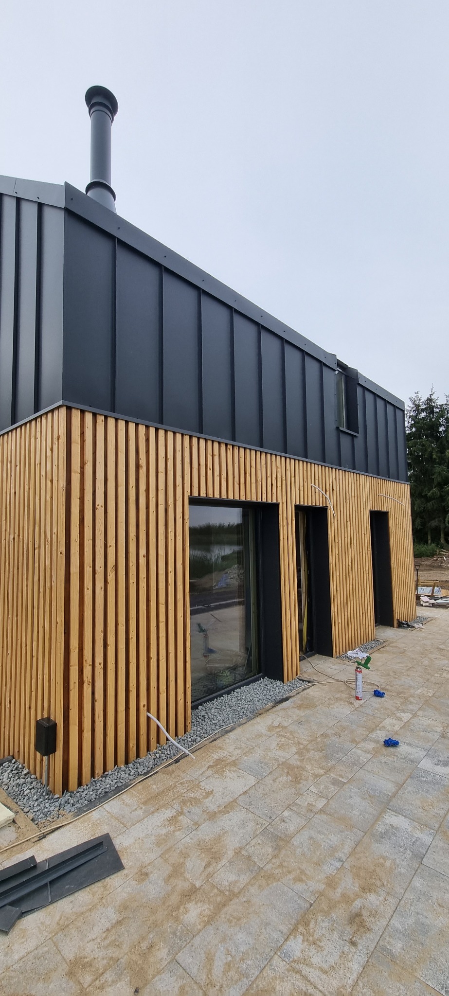 Nowoczesny dom z elewacją z pionowych desek i grafitowym dachem z blachy na rąbek stojący, widoczne okna z czarnymi ramami i fragment utwardzonego terenu z kostki brukowej.