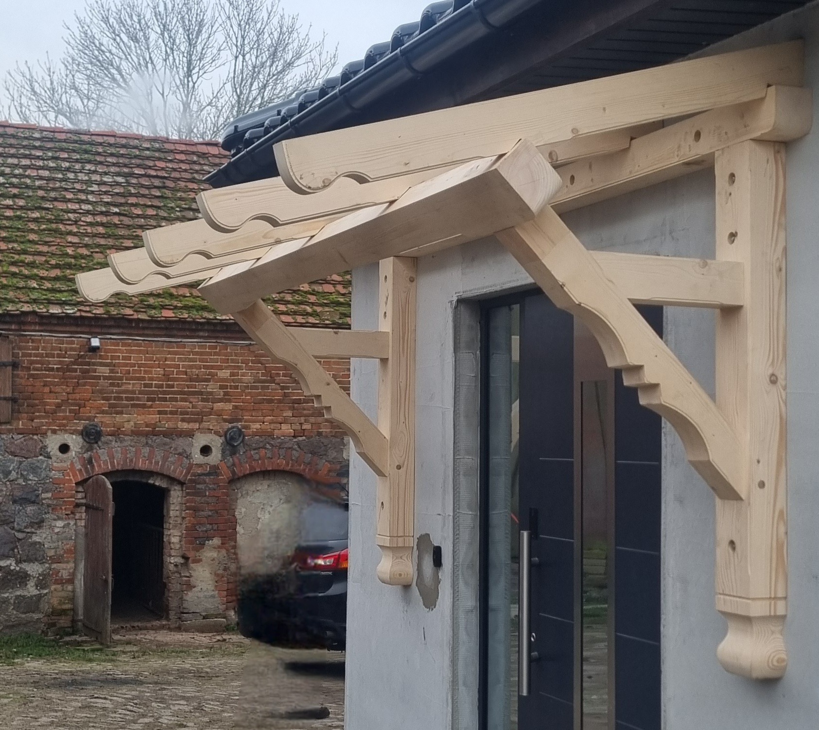 Drewniany, surowy daszek nad wejściem do budynku, z ozdobnymi wycięciami, w tle budynek z czerwonej cegły z otwartymi drzwiami.