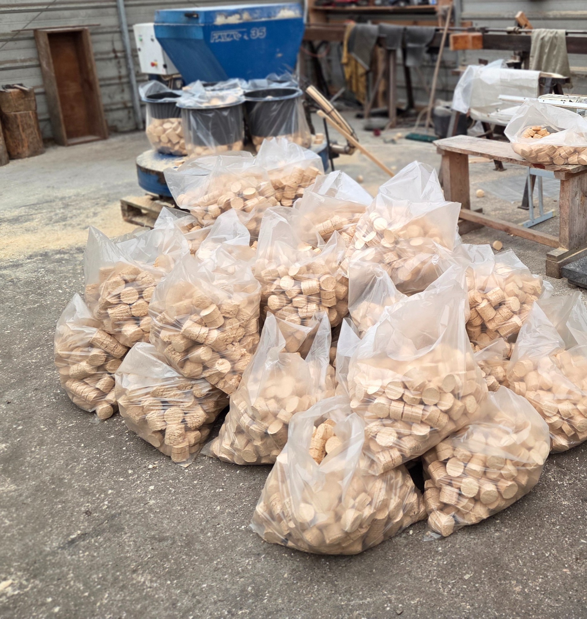 Stos przezroczystych worków wypełnionych drewnianymi pelletami, ustawionych na betonowej posadzce w hali produkcyjnej, z widocznym sprzętem do produkcji pelletu w tle.