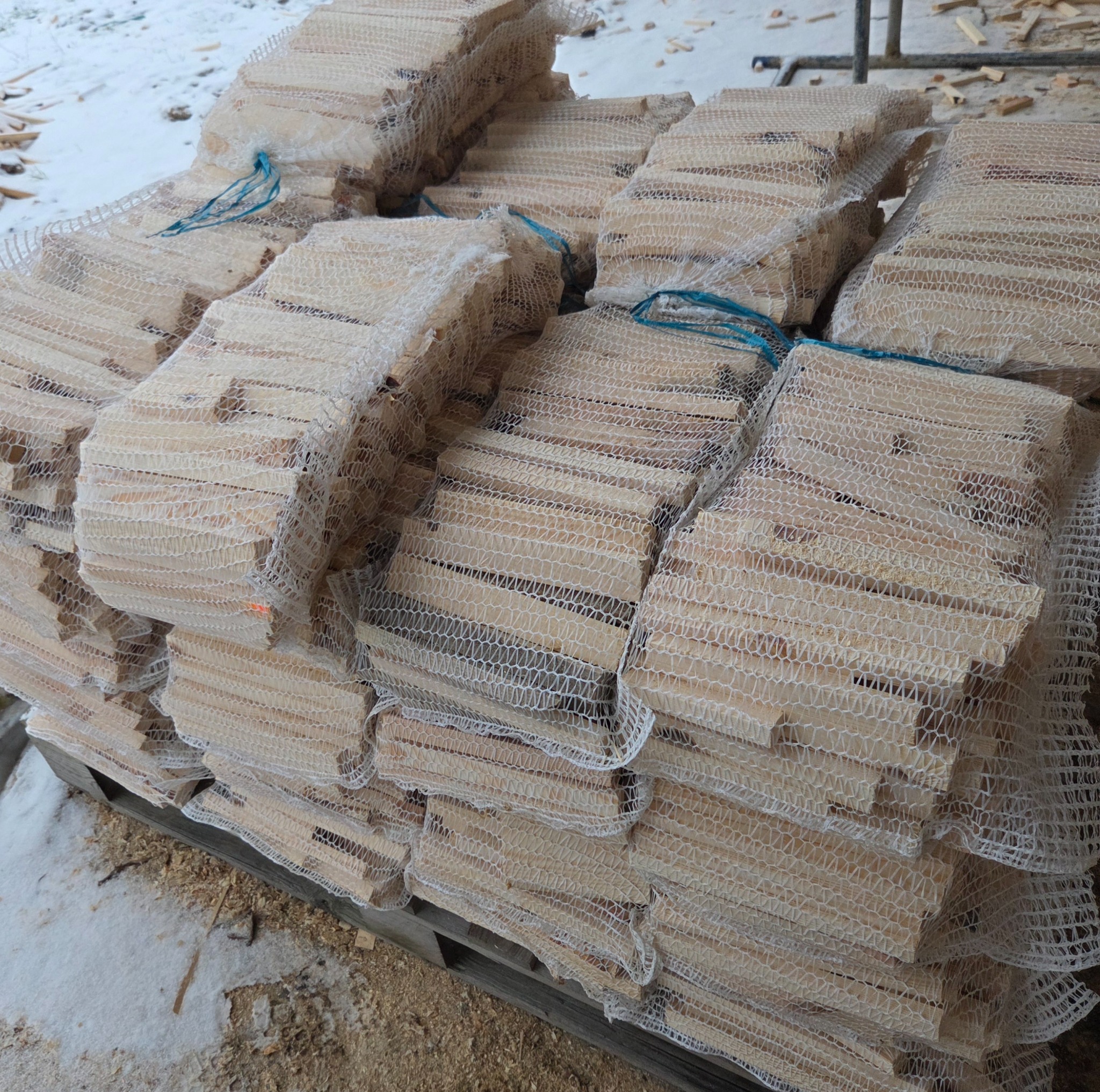 Paleta z workami wypełnionymi pociętym drewnem, zabezpieczonymi siatką z niebieskim sznurkiem, leżąca na śniegu i trocinach.