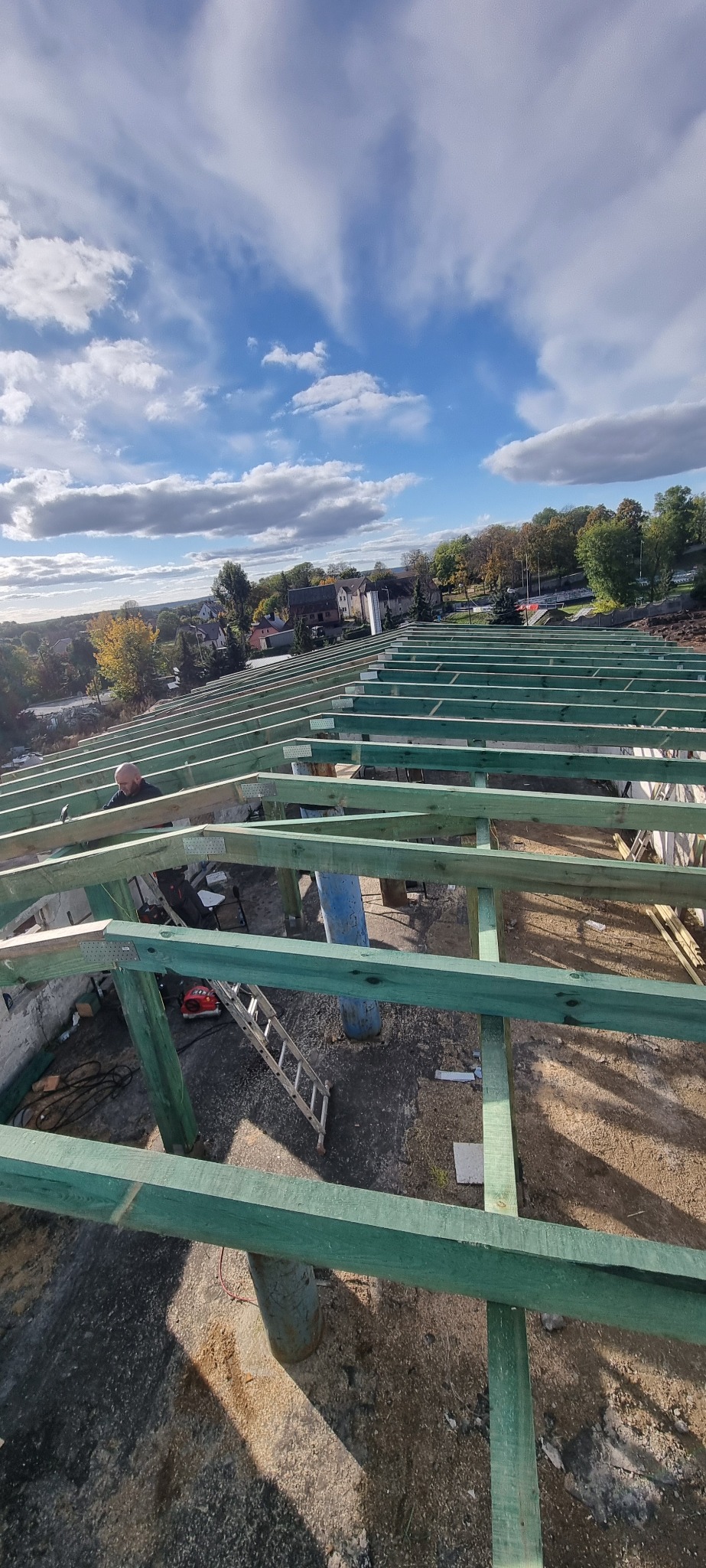 Konstrukcja dachu w trakcie budowy, widoczne zielone drewniane belki, drabina, narzędzia i pracownik w tle na tle błękitnego nieba z chmurami.