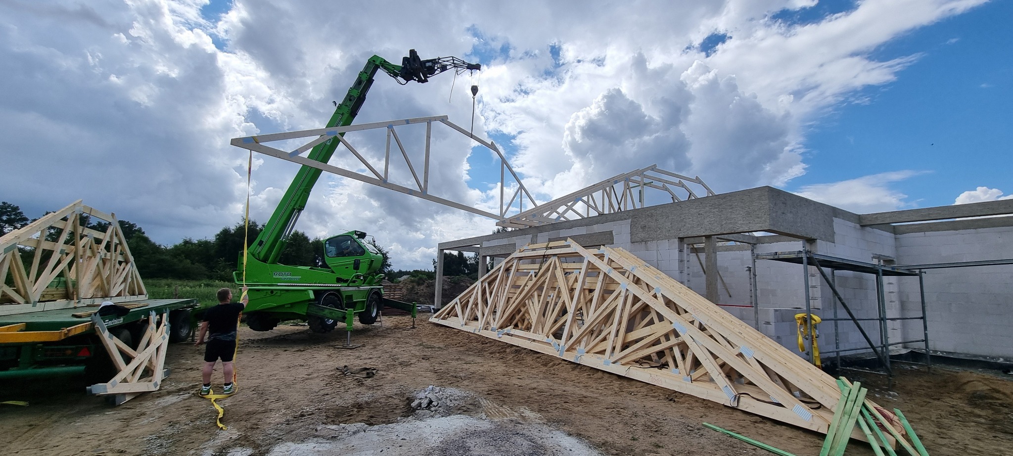 Montaż drewnianej więźby dachowej z użyciem dźwigu teleskopowego na placu budowy domu z białych bloczków, w tle widoczne drzewa i pochmurne niebo.