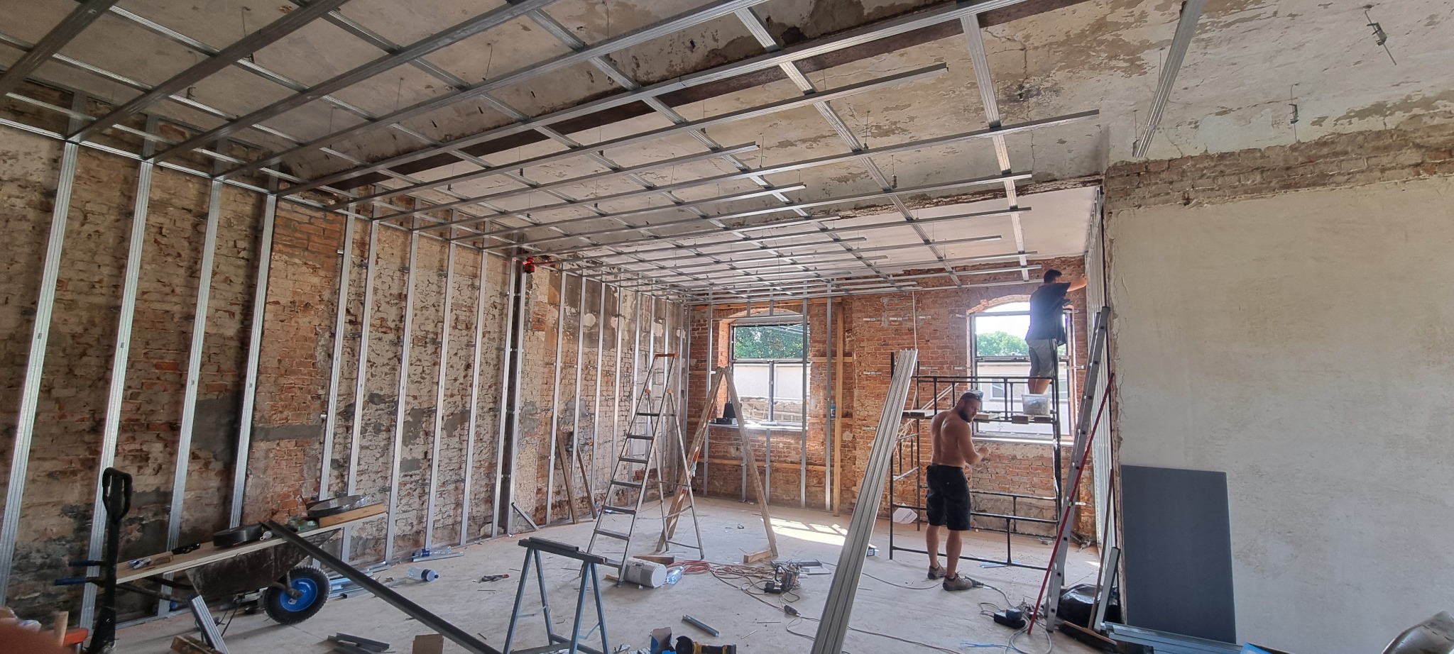 Montaż metalowej konstrukcji pod płyty gipsowo-kartonowe w pomieszczeniu z odsłoniętą cegłą, widoczne profile sufitowe i ścienne, dwaj pracownicy na drabinach, jeden bez koszulki.