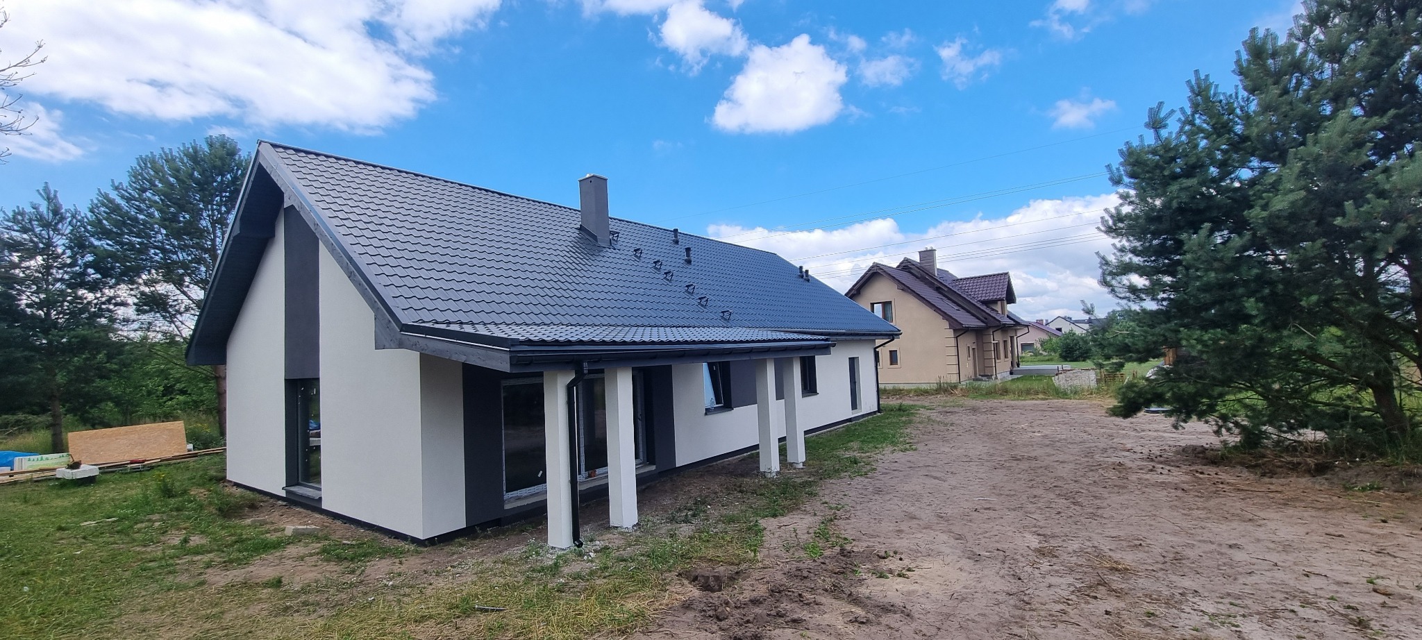 Nowoczesny dom z dwuspadowym dachem z blachy w kolorze grafitowym, biało-szara elewacja, widok z zewnątrz w słoneczny dzień, w tle inny dom i drzewa.