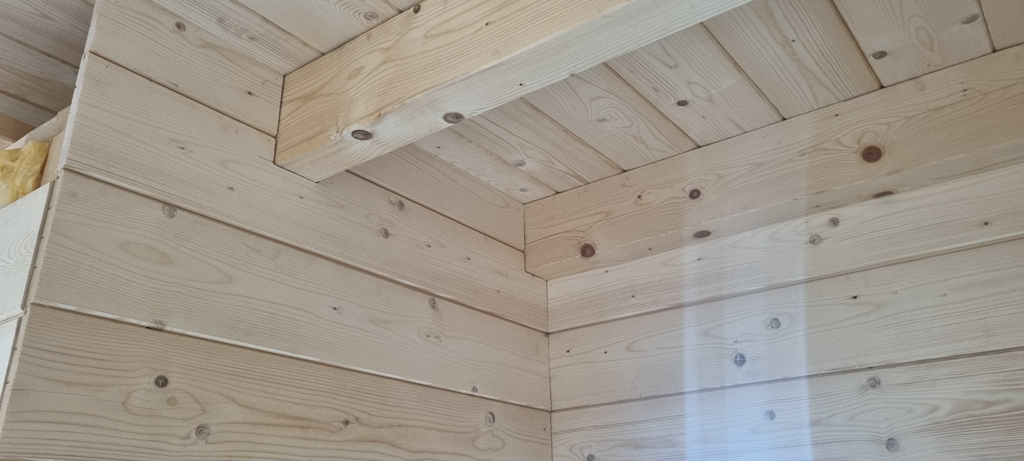 Wnętrze domu w trakcie budowy, widoczny fragment ściany i sufitu wykonane z jasnych, drewnianych desek łączonych na pióro i wpust, z widoczną izolacją z wełny mineralnej w narożniku.