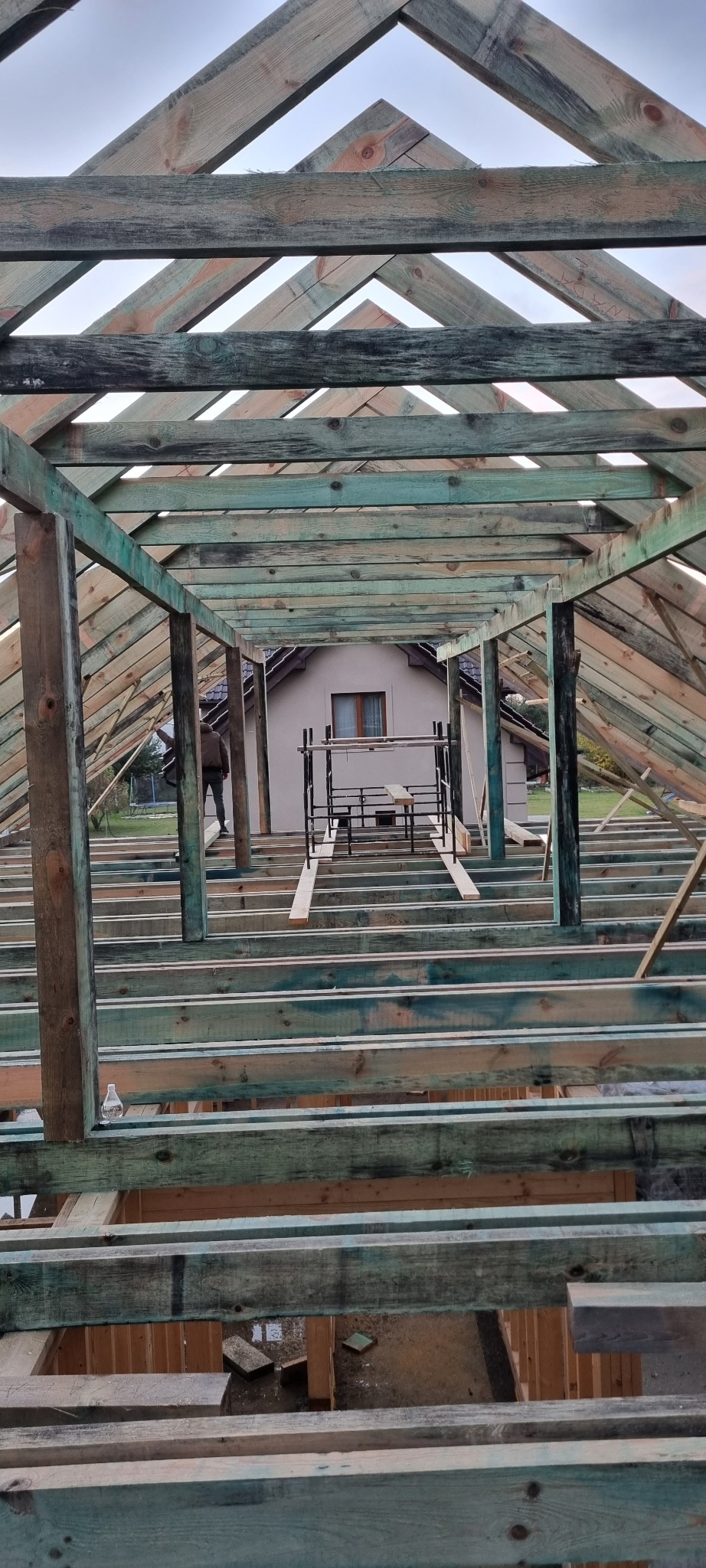 Konstrukcja drewniana dachu w trakcie budowy, widok od wewnątrz, z widocznym szkieletem i elementami więźby dachowej, w tle dom jednorodzinny i rusztowanie.