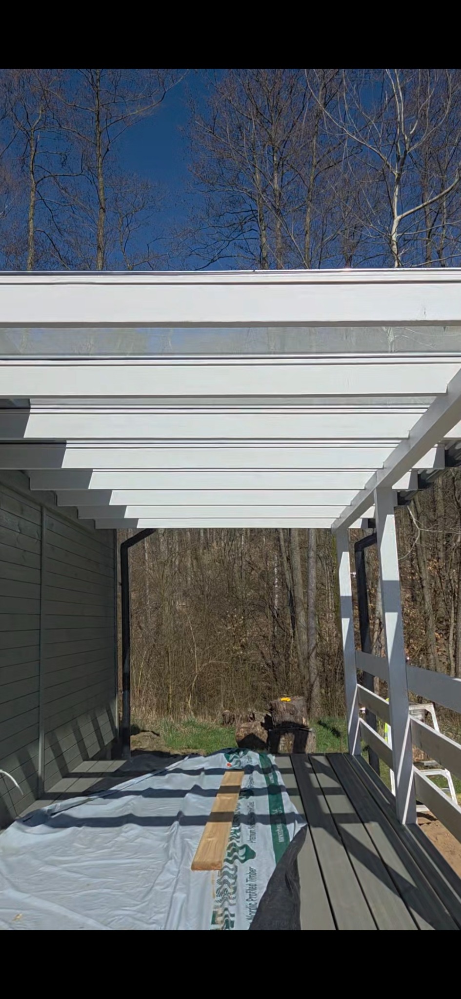 Biała konstrukcja zadaszenia tarasu w trakcie budowy, widoczna folia ochronna na podłodze tarasu i drewniane elementy przygotowane do montażu, w tle drzewa bez liści i błękitne niebo.