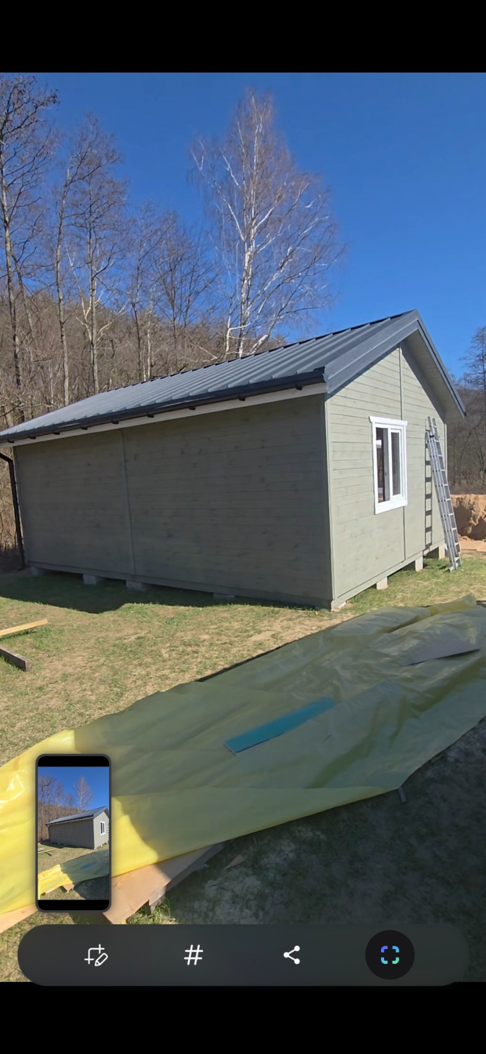 Niski, parterowy budynek z szarego drewna, widoczny z boku, z metalowym dachem i białą ramą okienną, stoi na trawie przykrytej żółtą folią budowlaną, z drabiną opartą o ścianę i drzewami w tle...